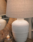 White ceramic table lamp with beaded detailing and linen lampshade on wooden table with small candle and mirror.