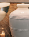 White ceramic table lamp with beaded detailing and linen lampshade on wooden table with small candle and mirror.