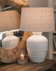 White ceramic table lamp with beaded detailing and linen lampshade on wooden table with small candle and mirror.