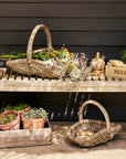 Two rattan trug baskets with plants on a wooden surface against a dark wall.