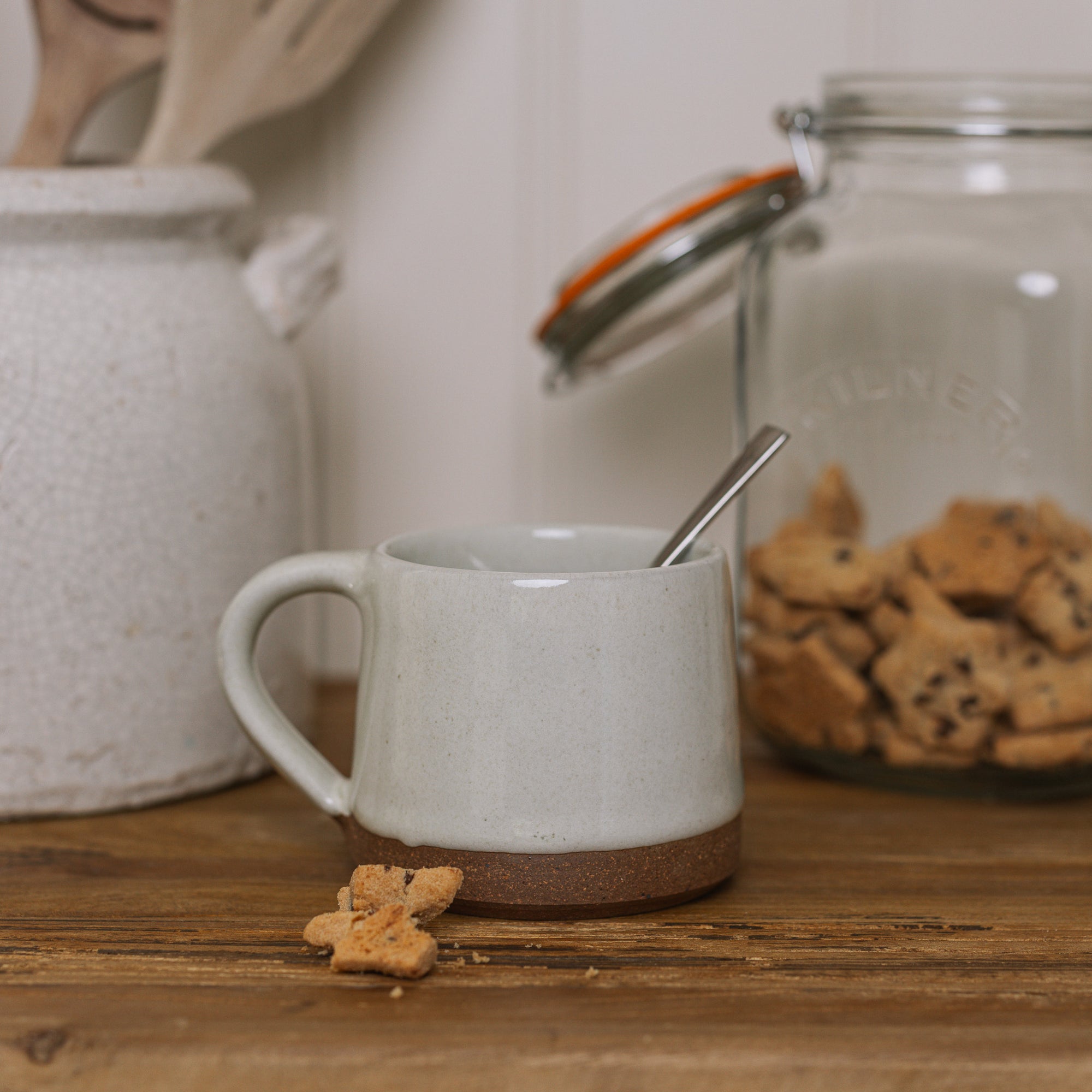 handmade milk white stoneware mug with stone base on wooden chopping board with crumbled biscuit.