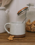 handmade milk white stoneware mug with stone base on wooden chopping board with crumbled biscuit.