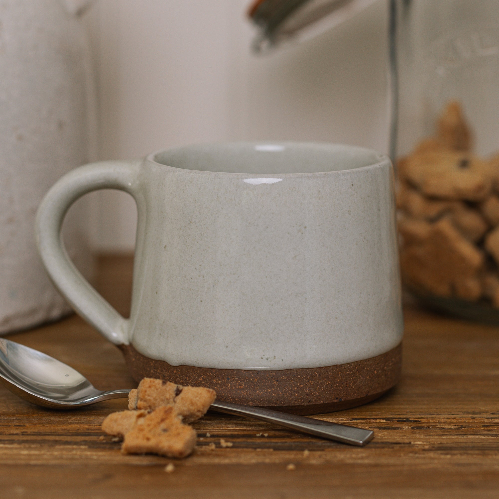 handmade milk white stoneware mug with stone base on wooden chopping board with crumbled biscuit.