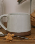 handmade milk white stoneware mug with stone base on wooden chopping board with crumbled biscuit.