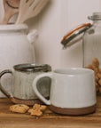 handmade milk white stoneware mug with stone base on wooden chopping board with crumbled biscuit.