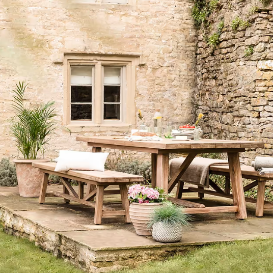 Large pine dining table outdoors with benches, plant pots and tableware.