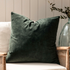 Square velvet cotton green cushion on rattan armchair with plant in vase behind.