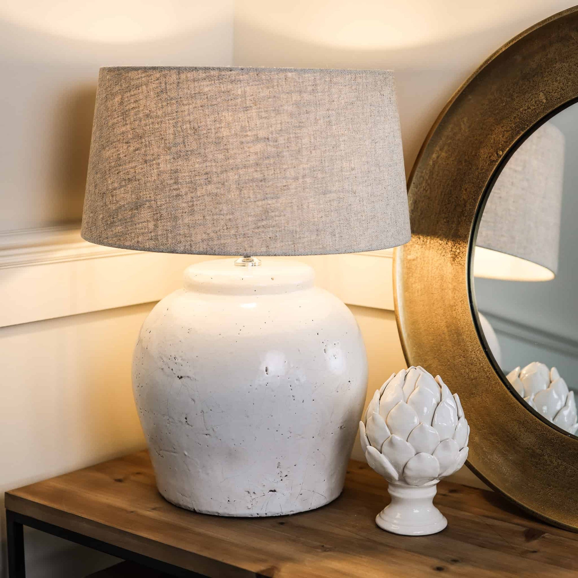 Distressed white lamp base with natural shade, switched on on wooden console with acorn ornament.