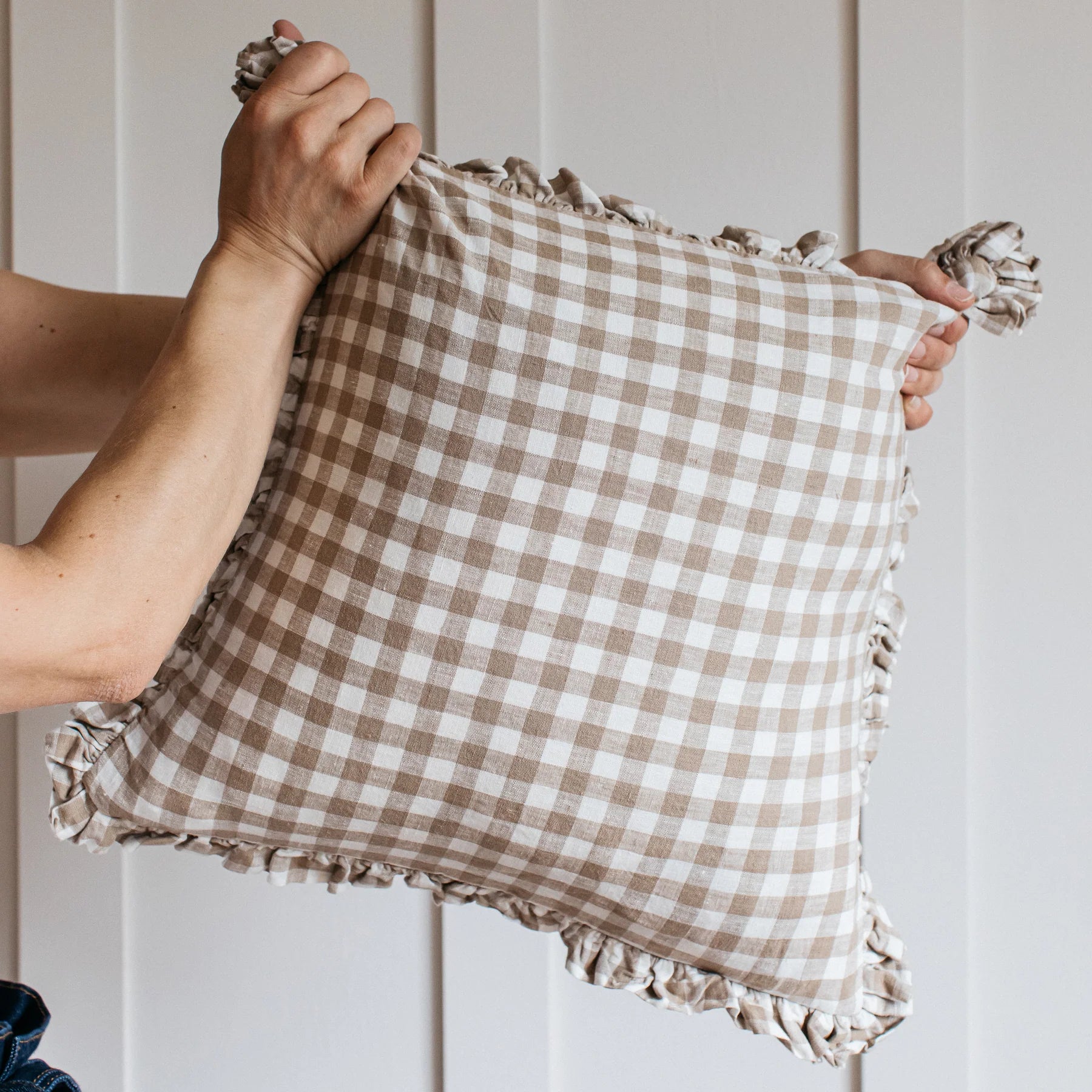 A person holds a neutral gingham ruffle cushion by each corner, there is a cream panelled wall in the background.