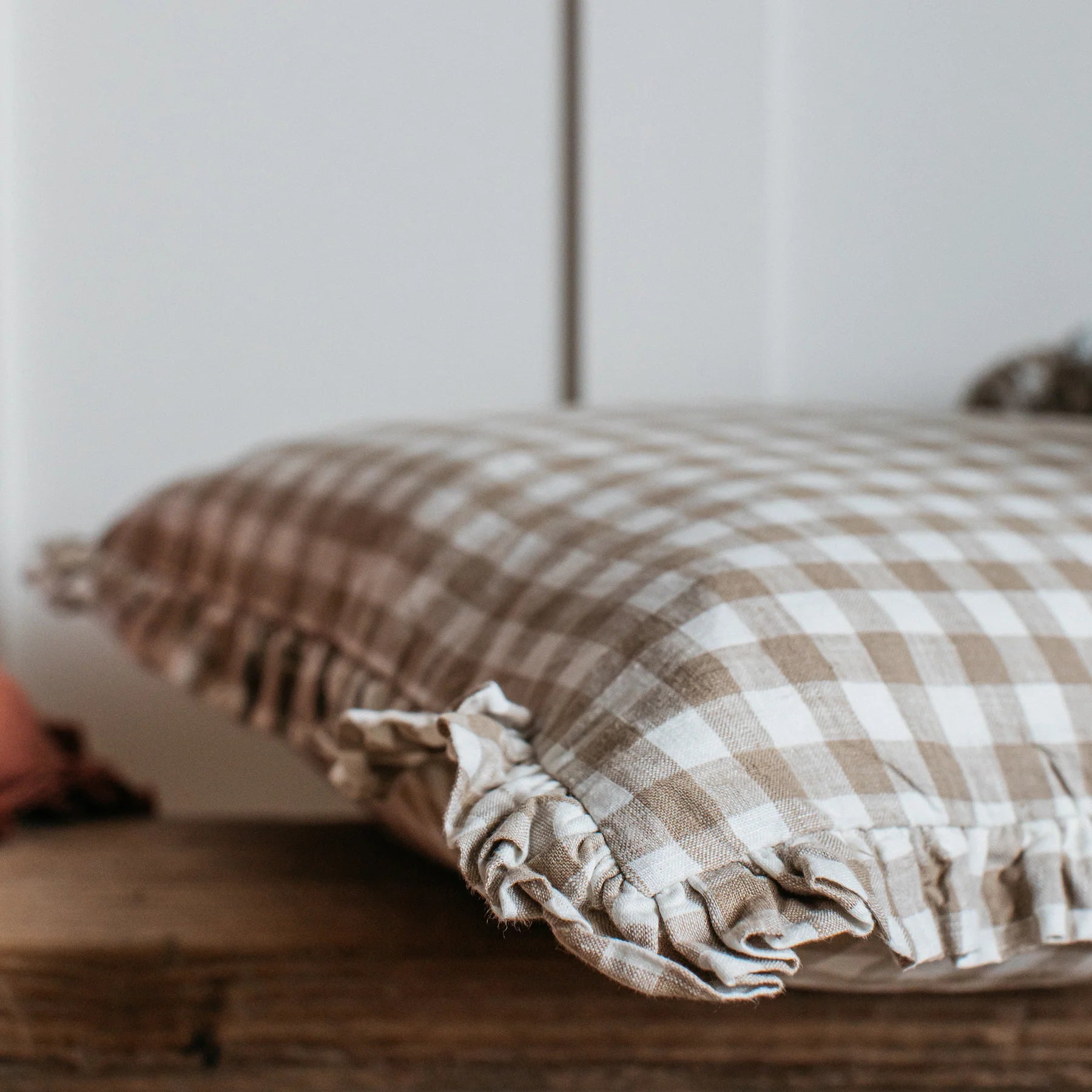 A neutral gingham ruffle cushion lays on a reclaimed style wooden bench against a cream panelled wall.