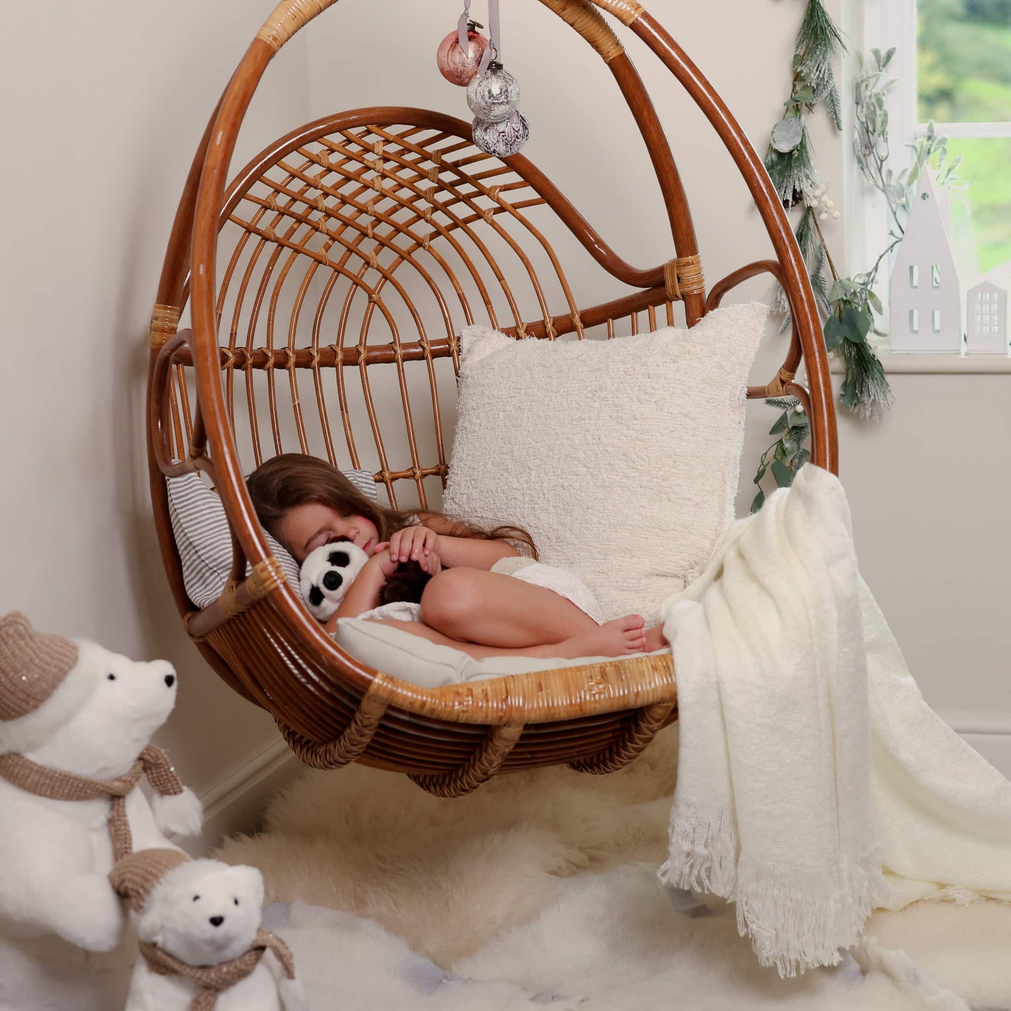 Natural rattan hanging chair for bedroom, with cushions and throw draped over and girl curled up with teddy.