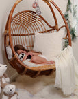 Natural rattan hanging chair for bedroom, with cushions and throw draped over and girl curled up with teddy.