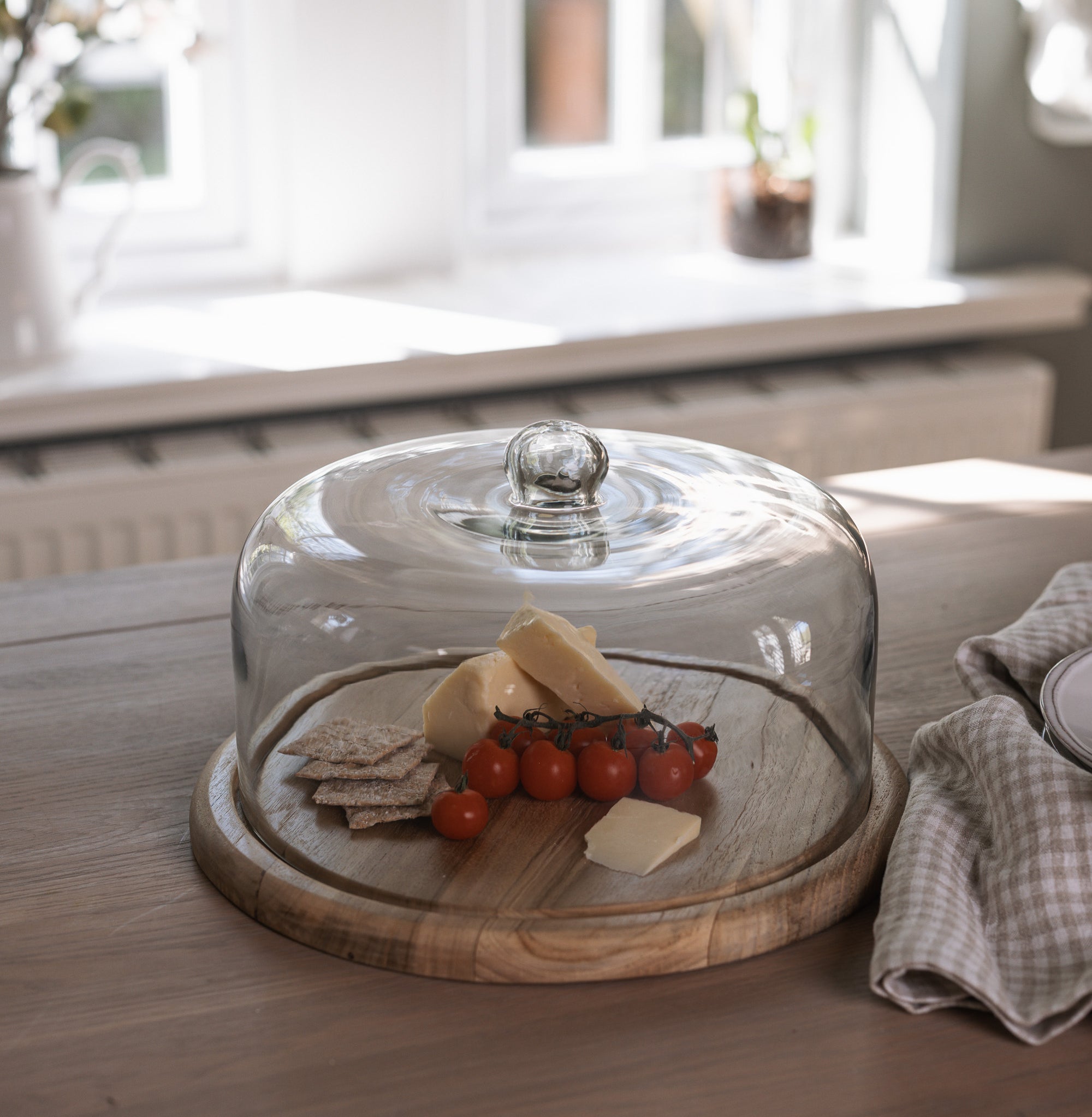 Recycled Glass Cake Dome filled with cheese, tomatoes and crackers with wooden base on wooden tale with gingham tea towel.