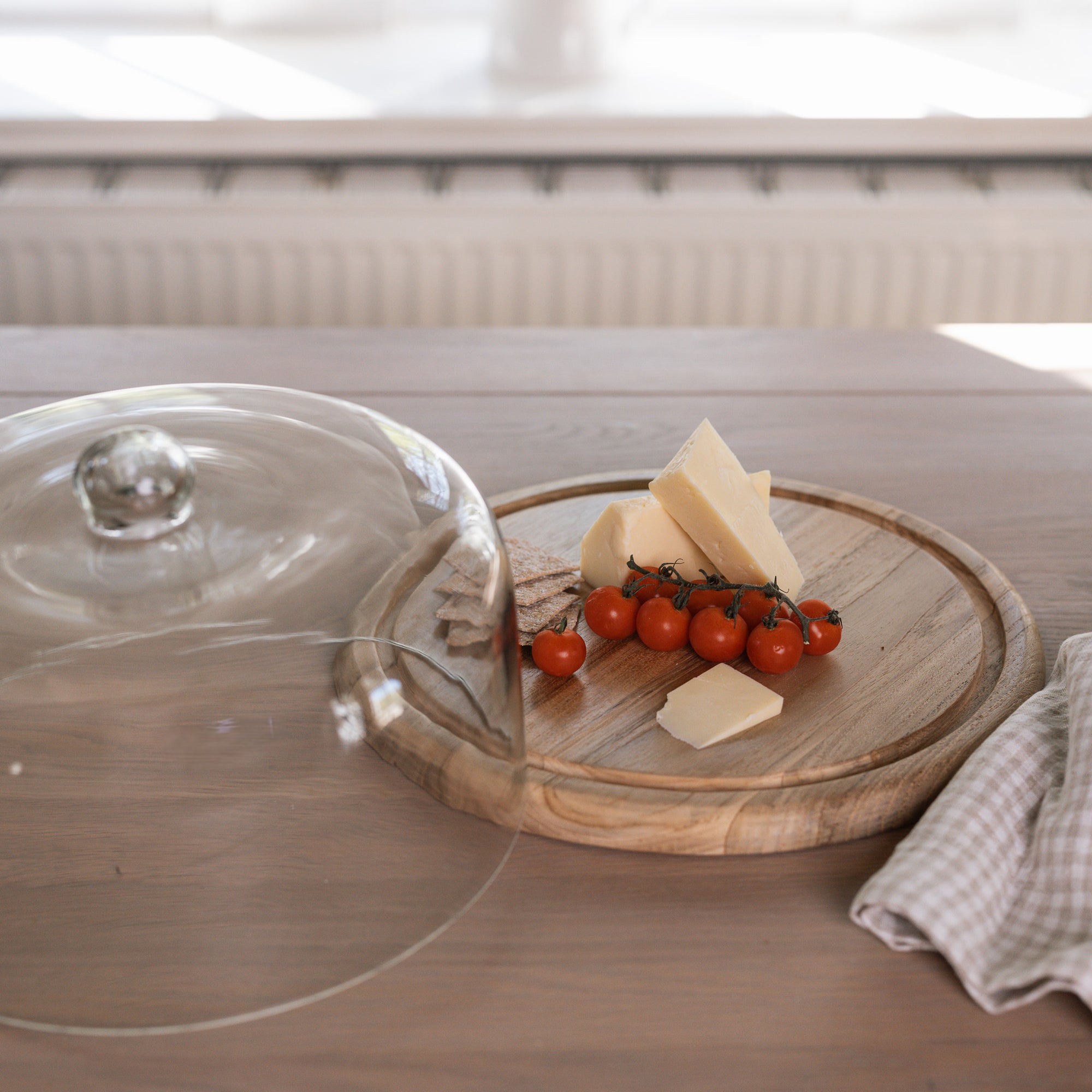 Recycled Glass Cake Dome filled with cheese, tomatoes and crackers with wooden base on wooden tale with gingham tea towel.