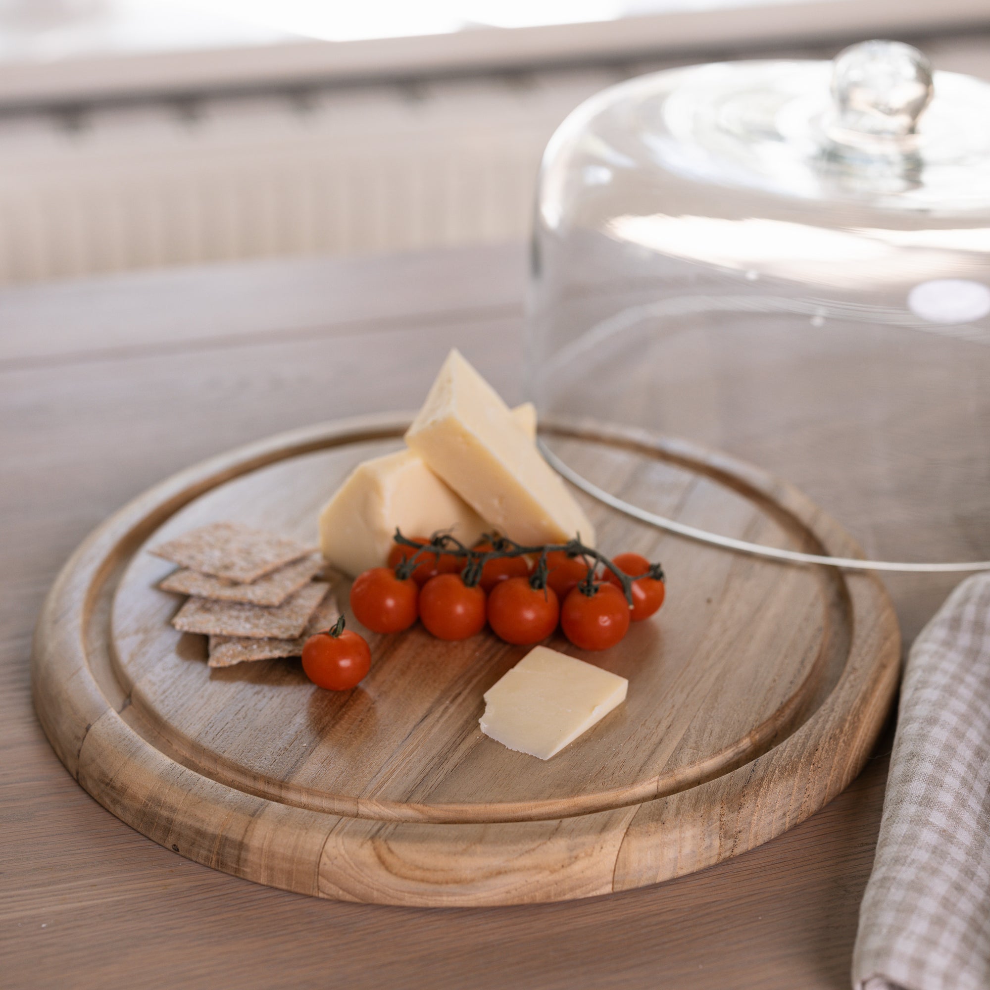 glass and wood cake dome with cheese, tomatoes and crackers on wooden table.