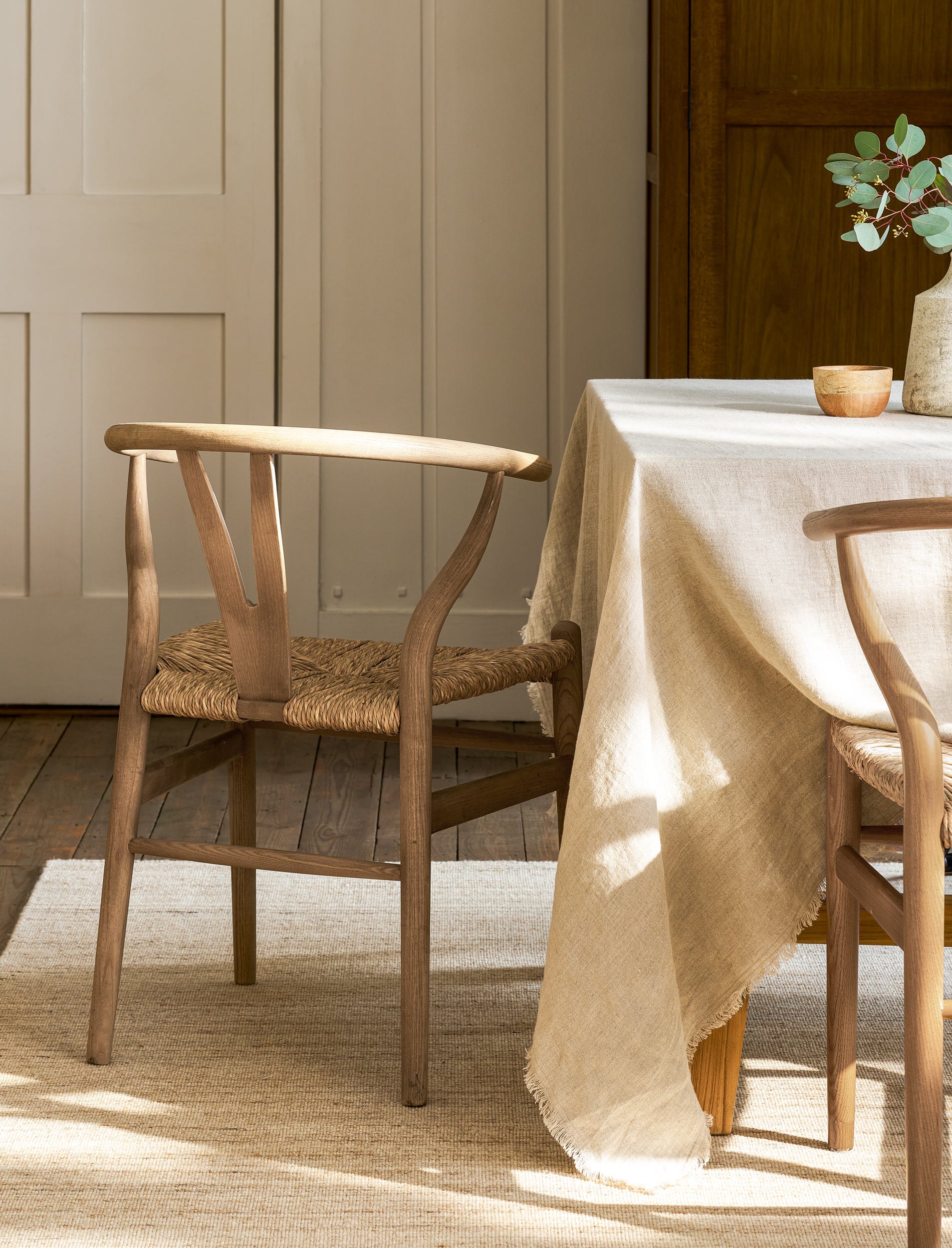 Wooden wishbone dining chairs with woven seat next to a table with a beige tablecloth in a room with wooden paneling.