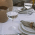 a textured wine glass filled with clear liquid on white linen tablecloth with lanterns in the background.