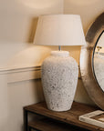 Large tall grey distressed lamp with white lamp shade, on wooden console.