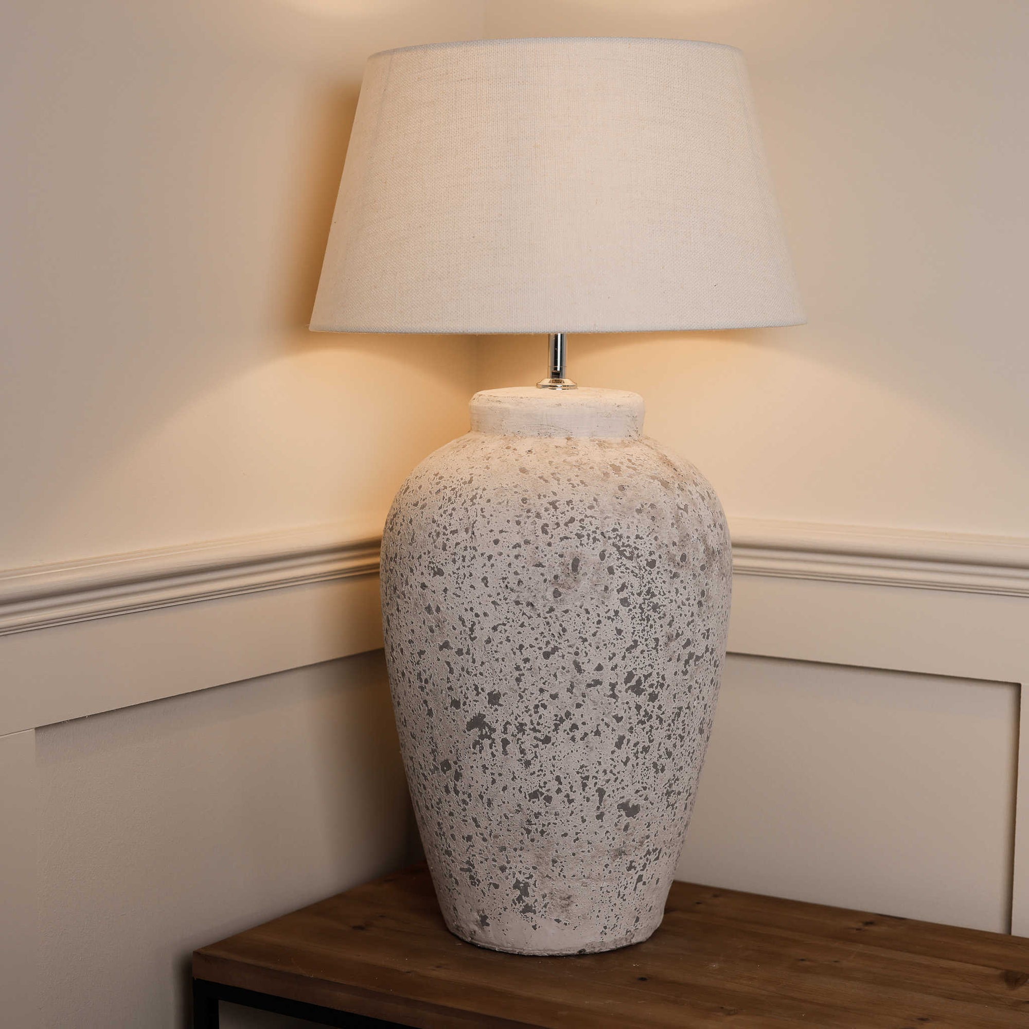 Large tall grey distressed lamp with white lamp shade, on wooden console, switched on.