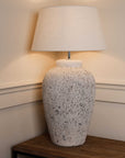 Large tall grey distressed lamp with white lamp shade, on wooden console, switched on.