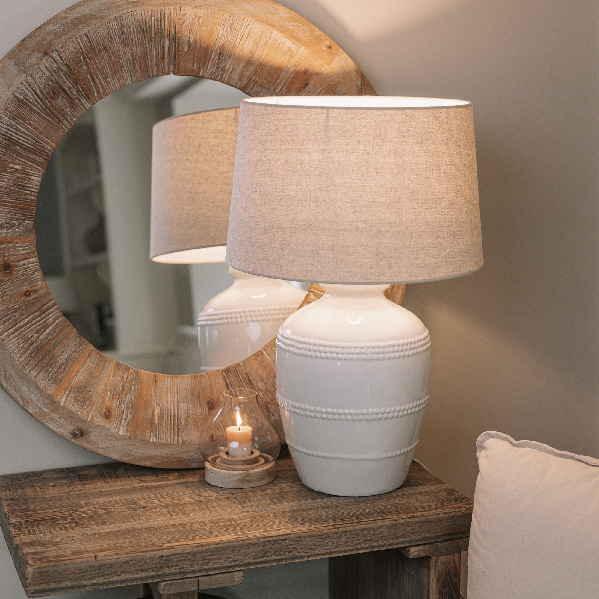 White ceramic table lamp with beaded detailing and linen lampshade on wooden table with small candle and mirror.