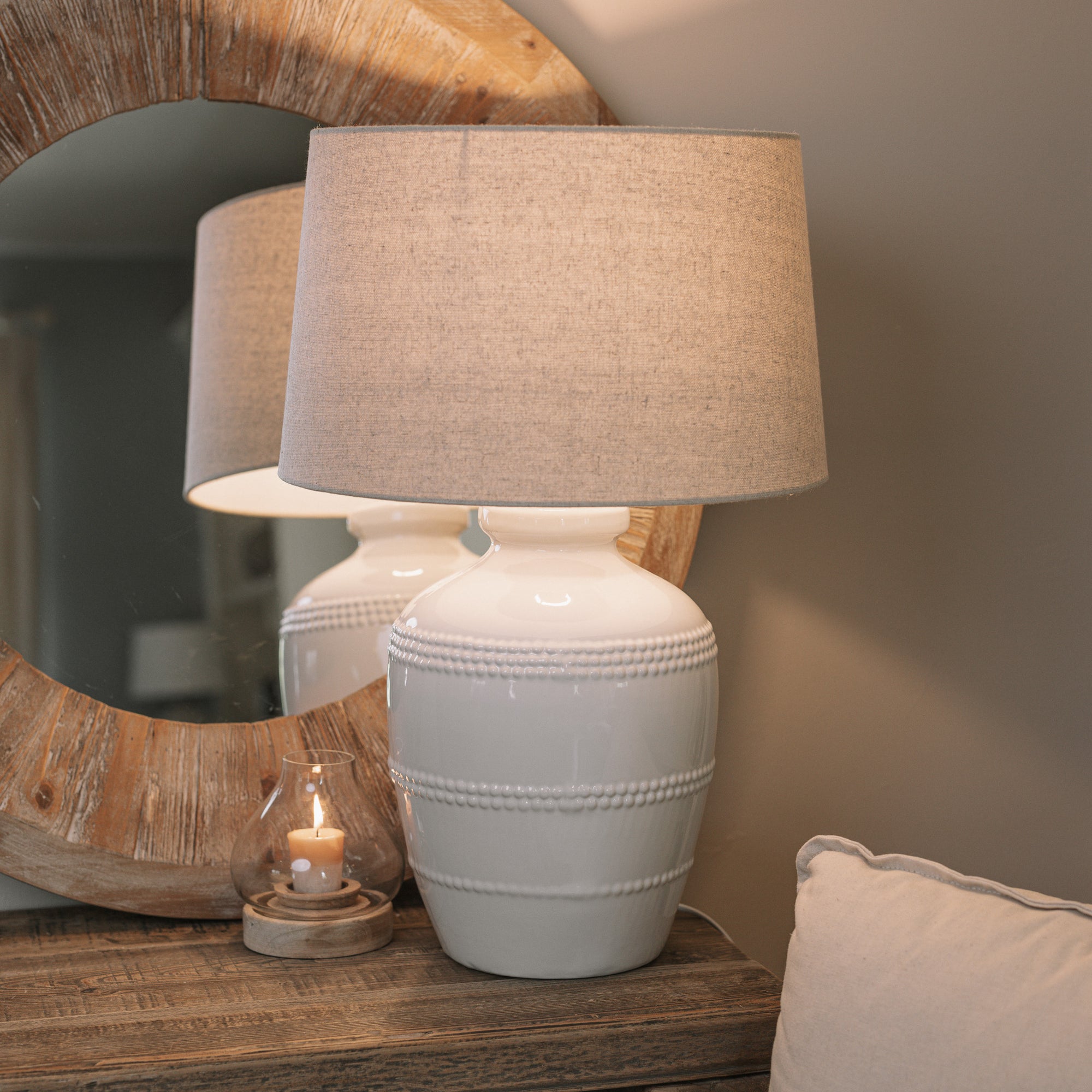 White ceramic table lamp with beaded detailing and linen lampshade on wooden table with small candle and mirror.