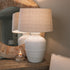 White ceramic table lamp with beaded detailing and linen lampshade on wooden table with small candle and mirror.