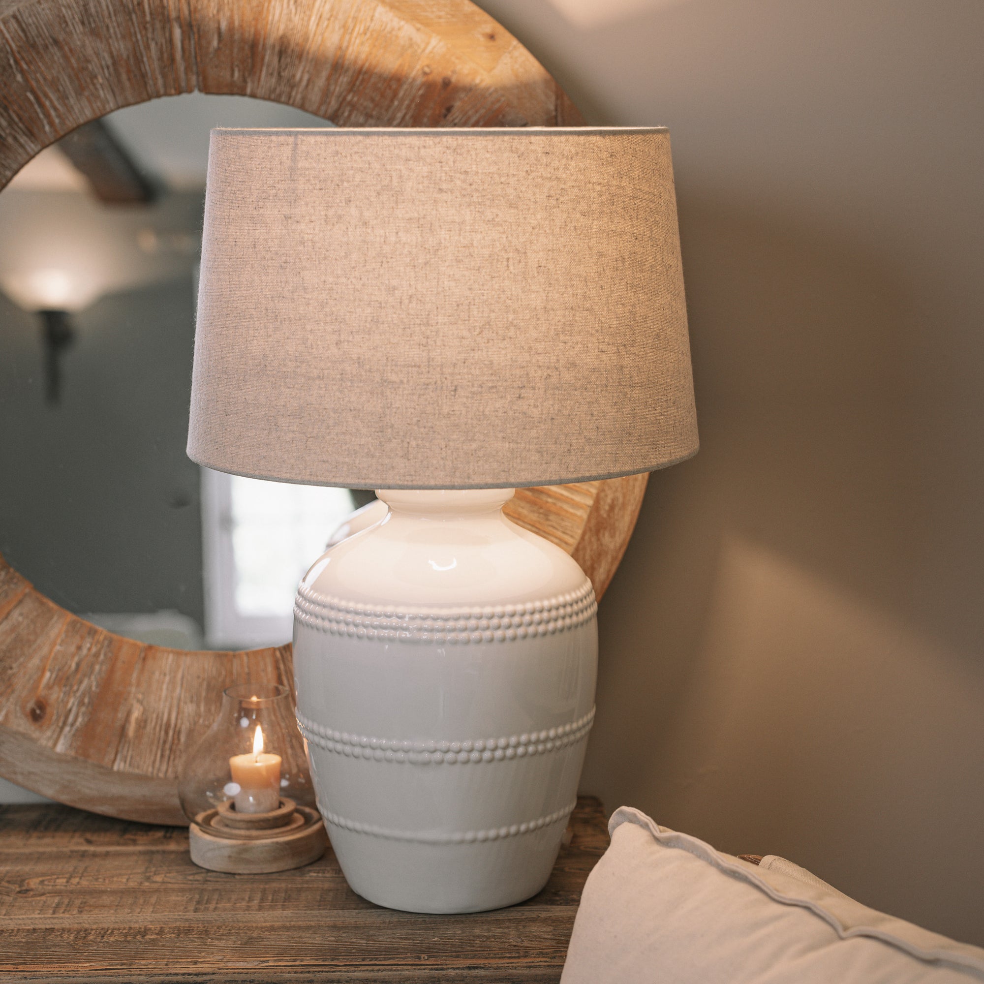 White ceramic table lamp with beaded detailing and linen lampshade on wooden table with small candle and mirror.
