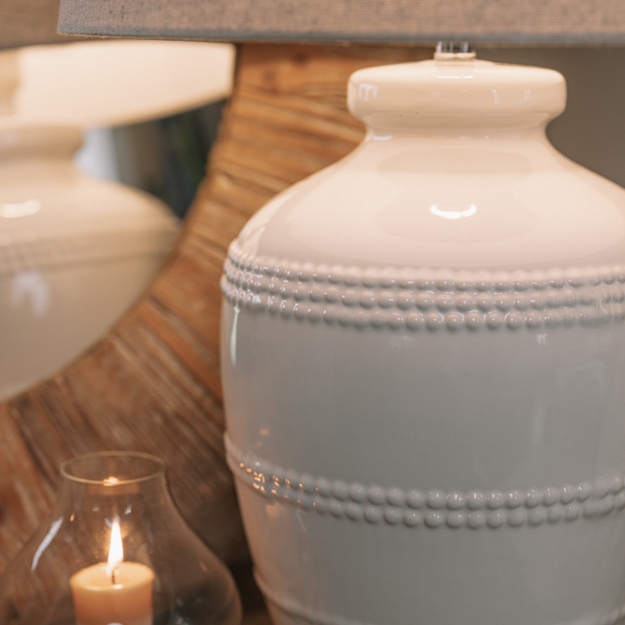 White ceramic table lamp with beaded detailing and linen lampshade on wooden table with small candle and mirror.