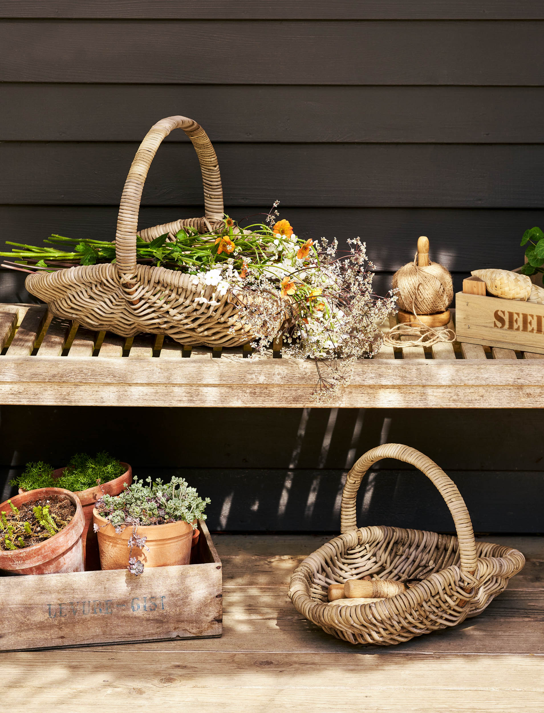 Two rattan trug baskets with plants on a wooden surface against a dark wall.