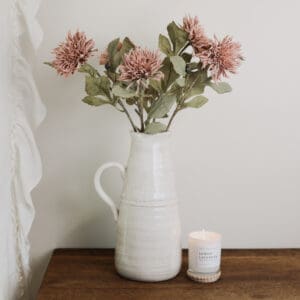 A white vase with pink Dahlia flowers and a small white candle, displayed on a wooden desk against a white wall.