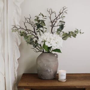 A green and white faux flower bouquet in a rustic grey vase on a wooden desk.