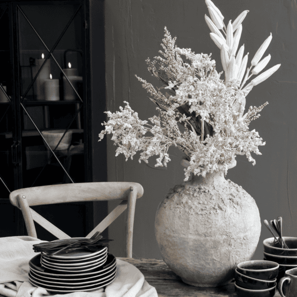 Rustic style vase with dried flowers, displayed on a dining table next to a stack of black plates.