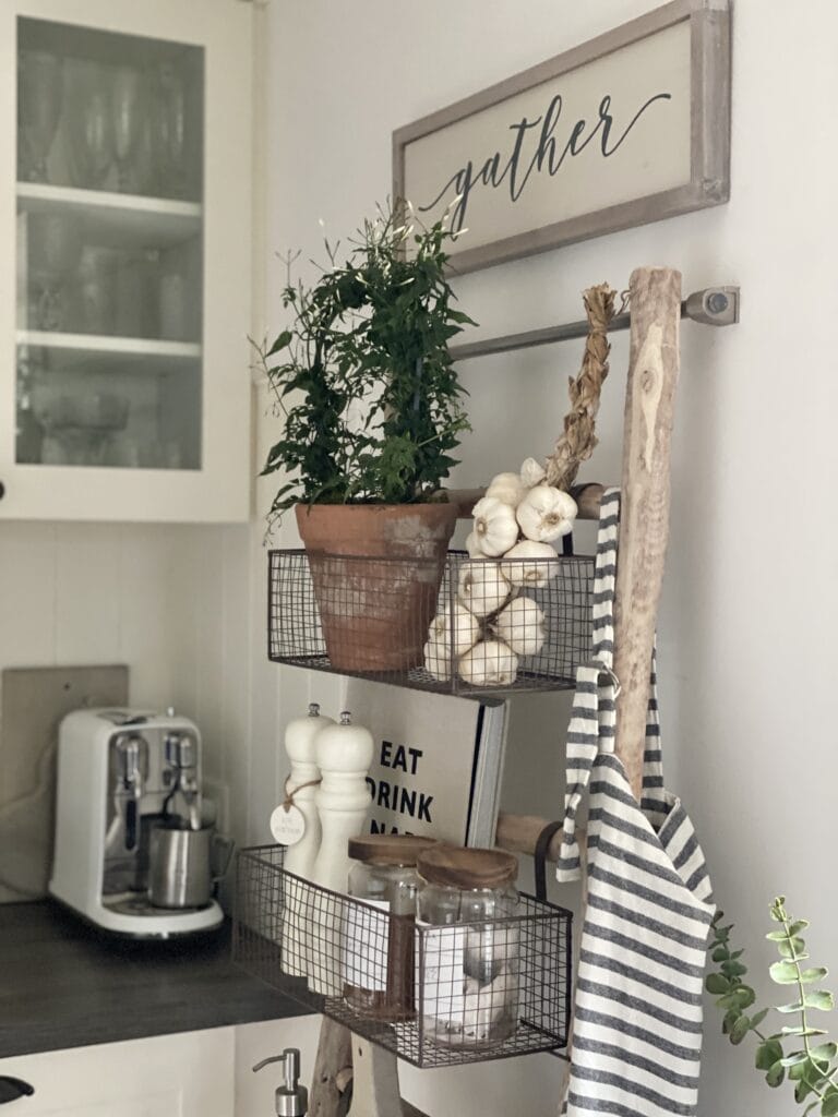 Wooden storage ladders with wire baskets in kitchen, filled with utensils, garlic and plants.