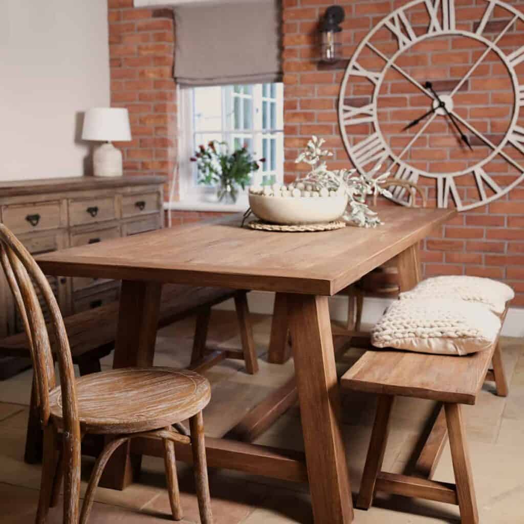 Farmhouse table and chairs in dining room, with cushions and brick wall.
